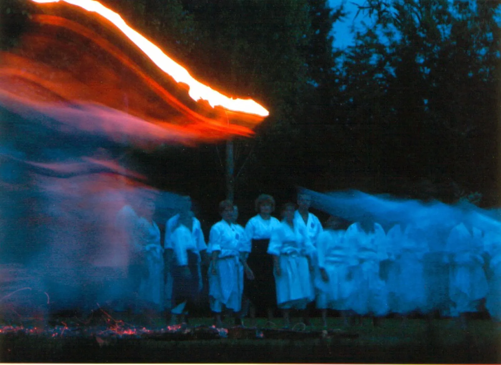 Nachtaufnahme mit Langzeitbelichtung: Eine Gruppe in weißen Kampfsport-Anzügen steht hinter einem Glutbett im Gras, verwischte Bewegungsspuren in Blau und ein orange leuchtender Flammenbogen ziehen durch das Bild, dahinter dunkle Bäume.