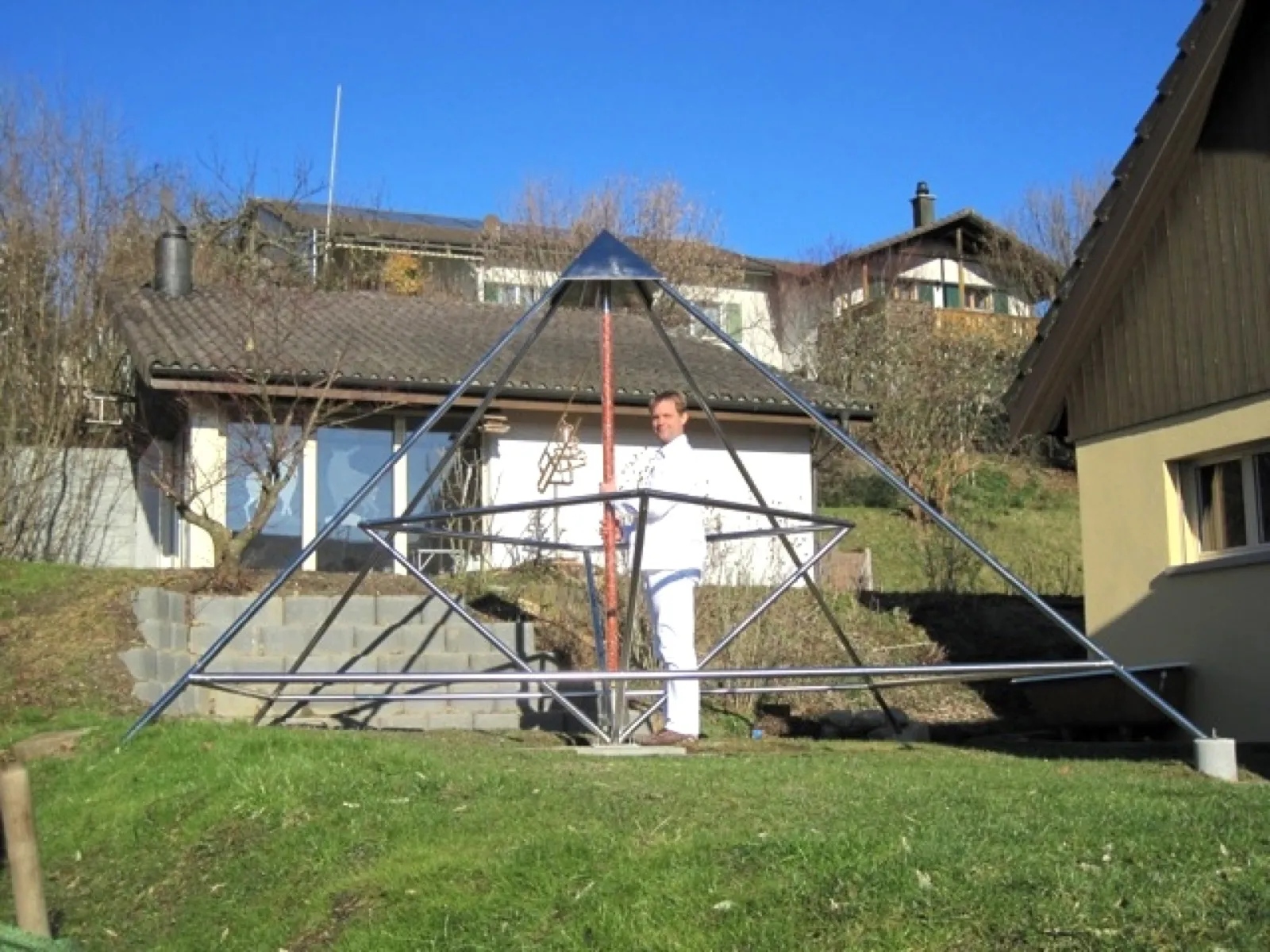 Ein Mann in weißer Kleidung steht in einer großen offenen Pyramide aus Metallstäben mit rot-weißer Mittelachse und glänzender Spitze auf einer Wiese; dahinter Wohnhäuser am Hang unter blauem Himmel.