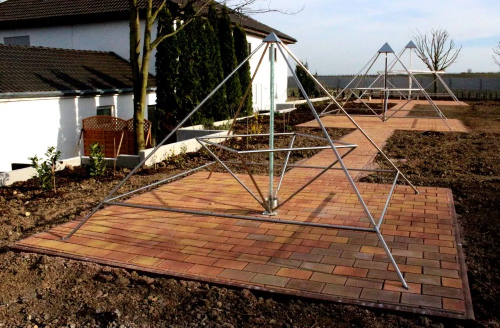 Eine große offene Edelstahlpyramide mit verdrehter innerer Pyramide steht auf einer rot-gelb gepflasterten Fläche inmitten frisch umgegrabener Erde; ein gepflasterter Weg führt zu zwei weiteren Pyramiden. Links ein weißes Wohnhaus, Zypressen und ein junger Baum.