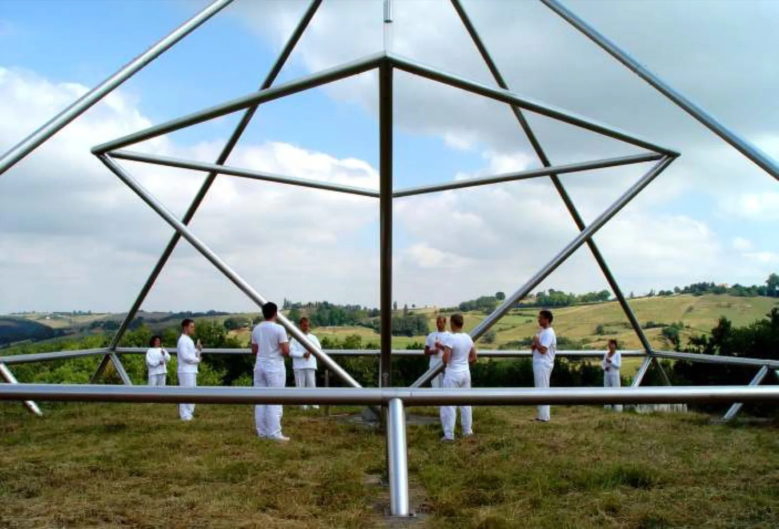 Blick aus dem Inneren einer großen Pyramidenanlage aus Edelstahlrohren: Etwa ein Dutzend Menschen in weißer Kleidung steht im Kreis auf der Wiese, dahinter eine sommerliche Hügellandschaft unter Wolken.