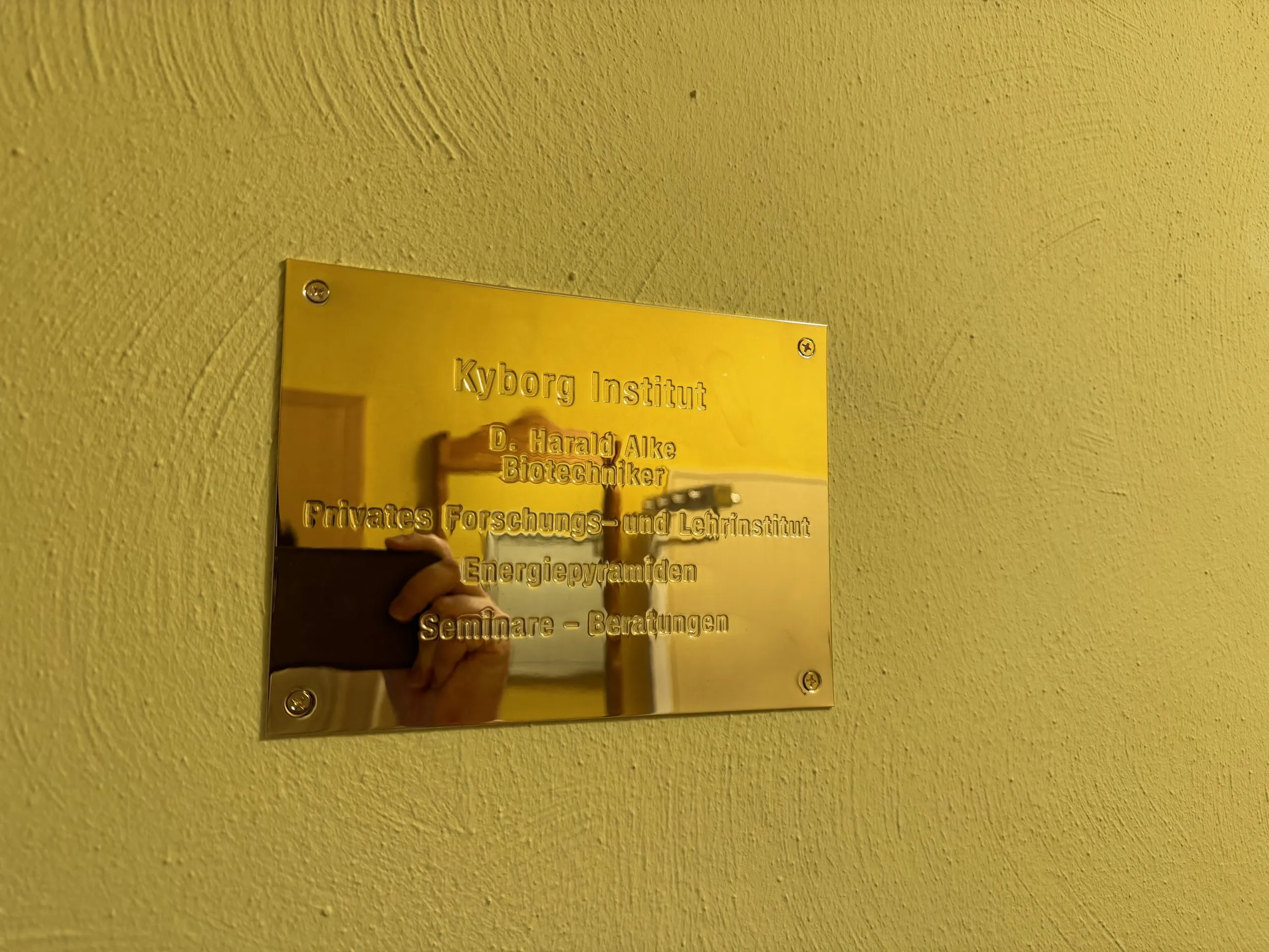Messing-Schild an gelber Wand mit der Aufschrift „Kyborg Institut – D. Harald Alke, Biotechniker – Privates Forschungs- und Lehrinstitut – Energiepyramiden – Seminare, Beratungen“.