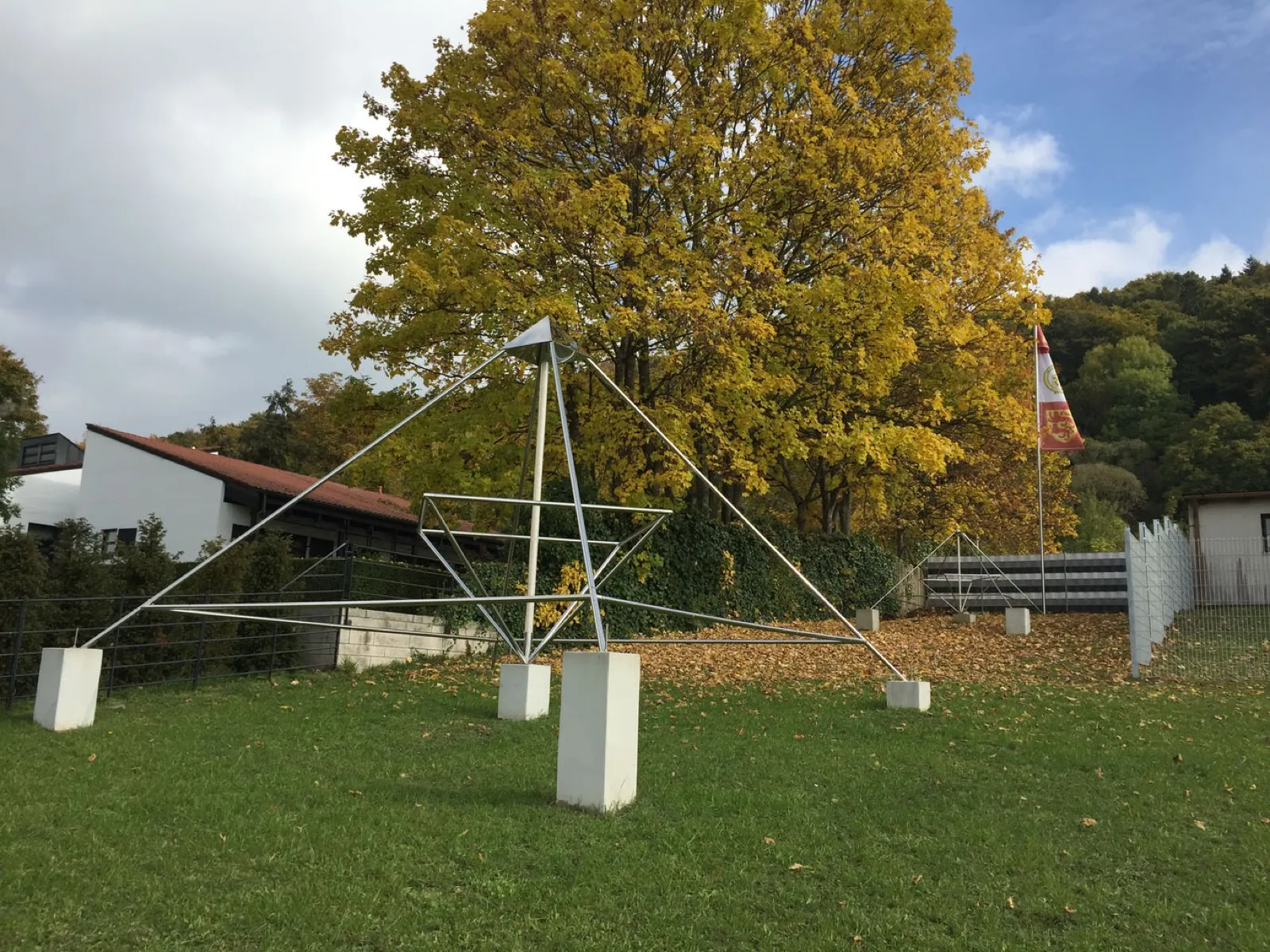 Große offene Pyramide aus Edelstahlrohren auf weißen Betonsockeln auf einer Rasenfläche vor einem gelb gefärbten Herbstbaum; rechts dahinter eine kleinere Pyramide auf Laub, ein Fahnenmast mit rot-weißer Löwenfahne und ein weißes Gebäude mit rotem Dach.