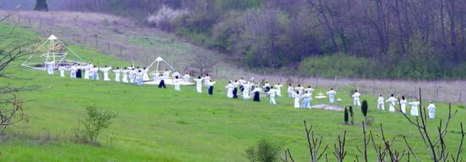 Eine große Gruppe weiß gekleideter Menschen übt verteilt auf einer grünen Hangwiese mit ausgebreiteten Armen; links stehen zwei offene Pyramidengestelle aus Metall, im Hintergrund Gebüsch und kahle Bäume.