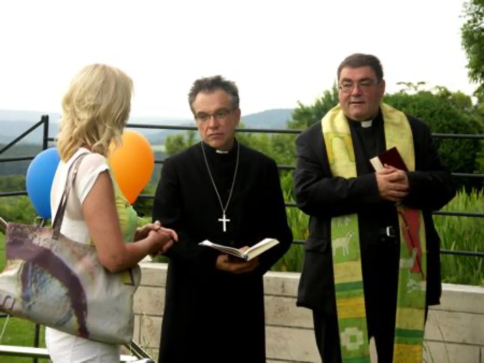 Zwei Geistliche stehen im Freien vor einem Geländer mit Blick ins Grüne: links einer in schwarzem Talar mit Brustkreuz und aufgeschlagenem Buch, rechts einer mit grüner, bestickter Stola und Buch in der Hand. Vorne links eine blonde Frau von hinten mit Umhängetasche, daneben blaue und orange Luftballons.