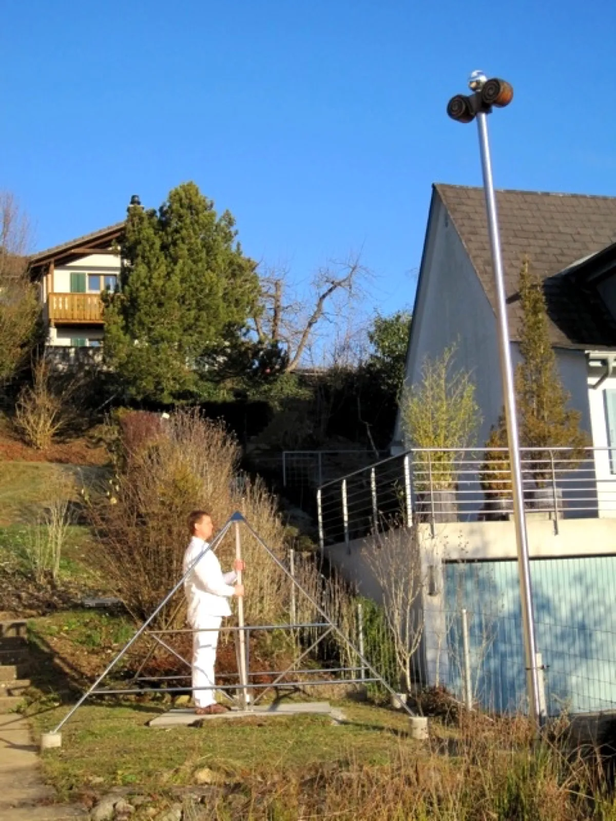 Ein Mann in weißer Kleidung steht in einer offenen Pyramide aus Metallstäben auf einem Gartenhang und hält den mittigen Stab; daneben ein hoher Mast mit zwei Strahlern, im Hintergrund Wohnhäuser unter blauem Himmel.