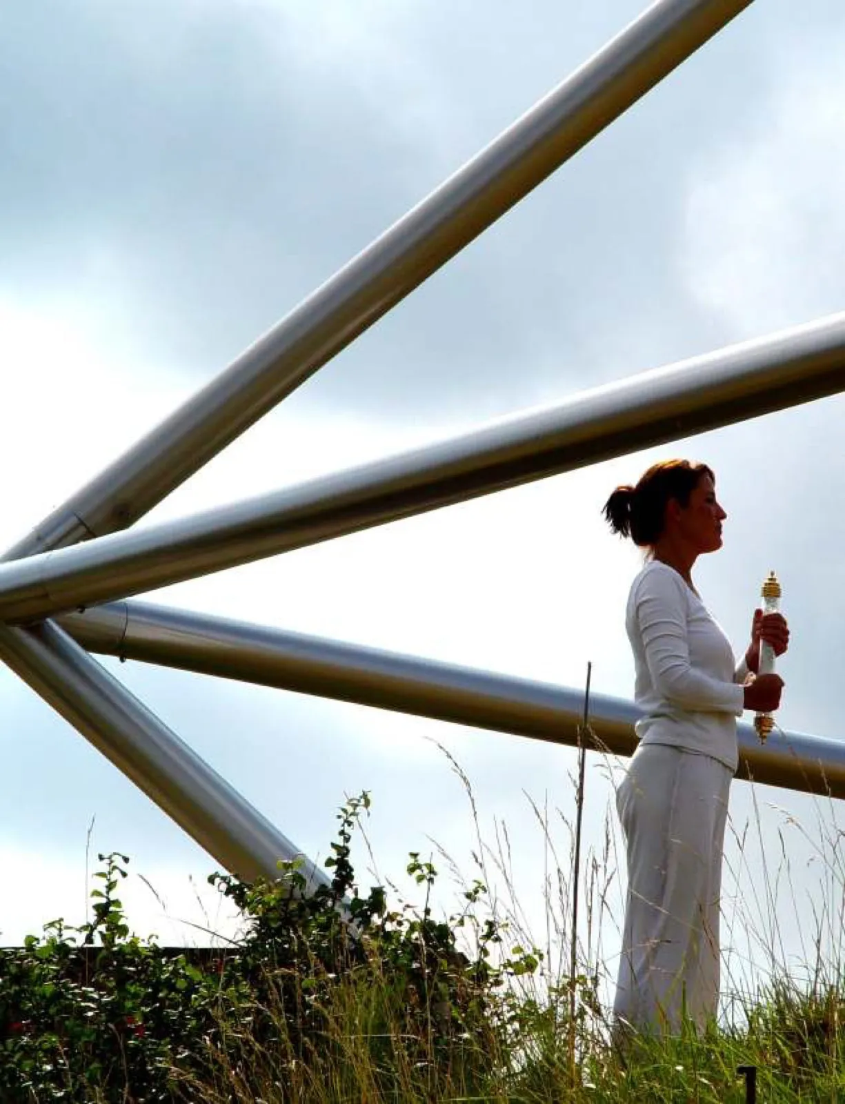 Eine Frau in weißer Kleidung steht im hohen Gras vor den schräg verlaufenden Edelstahlrohren einer großen Pyramidenanlage und hält einen Stab mit Glaskörper und goldenen Endkappen; im Hintergrund bewölkter Himmel.