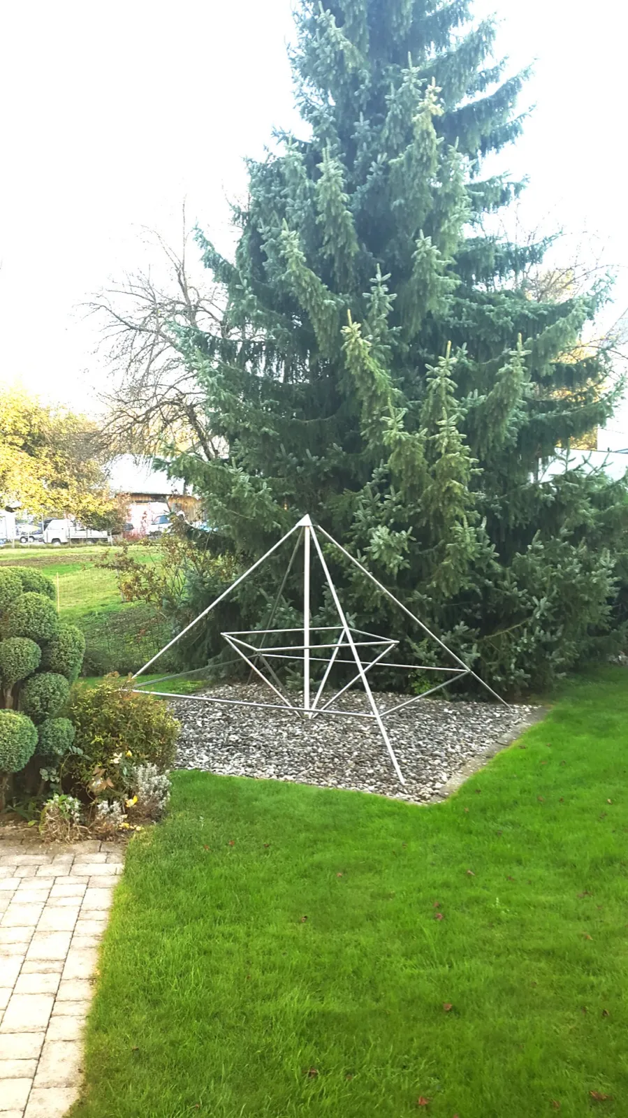 Eine offene Edelstahl-Energiepyramide mit innerer verdrehter Pyramide steht auf einem Kiesbett vor einer hohen Fichte in einem Garten mit gepflegtem Rasen, Kugelbuchs und Pflasterweg. Im Hintergrund eine Wiese, eine Scheune und abgestellte Fahrzeuge.