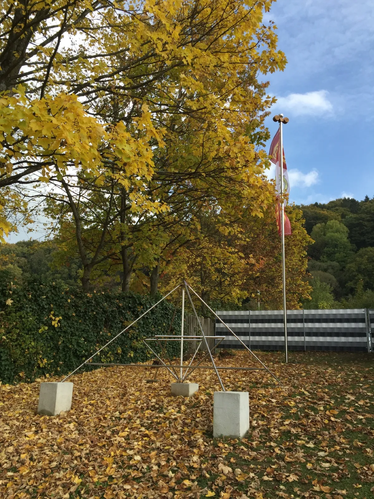 Herbstlicher Garten mit gelb gefärbten Bäumen und Laub am Boden: vorne eine offene Edelstahlpyramide auf Betonsockeln, daneben ein hoher Mast mit Spiralvoluten-Kapitell und rot-weißer Fahne mit Emblem, dahinter Efeuhecke, gestreifter Zaun und bewaldeter Hang.