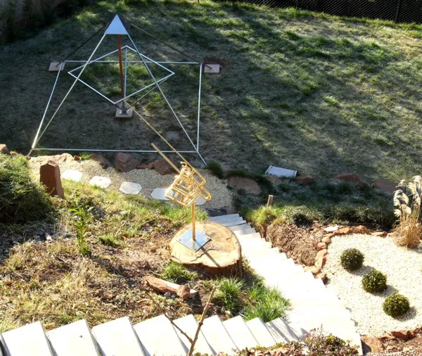 Große offene Energiepyramide aus Edelstahlstäben mit kupferfarbener Mittelachse auf Steinplatten im Rasen; im Inneren steht eine Person in blauer Jacke und dunkler Mütze an der Mittelachse. Dahinter eine Sandsteinmauer, ein Schilfrohrzaun und hohe Nadelbäume.
