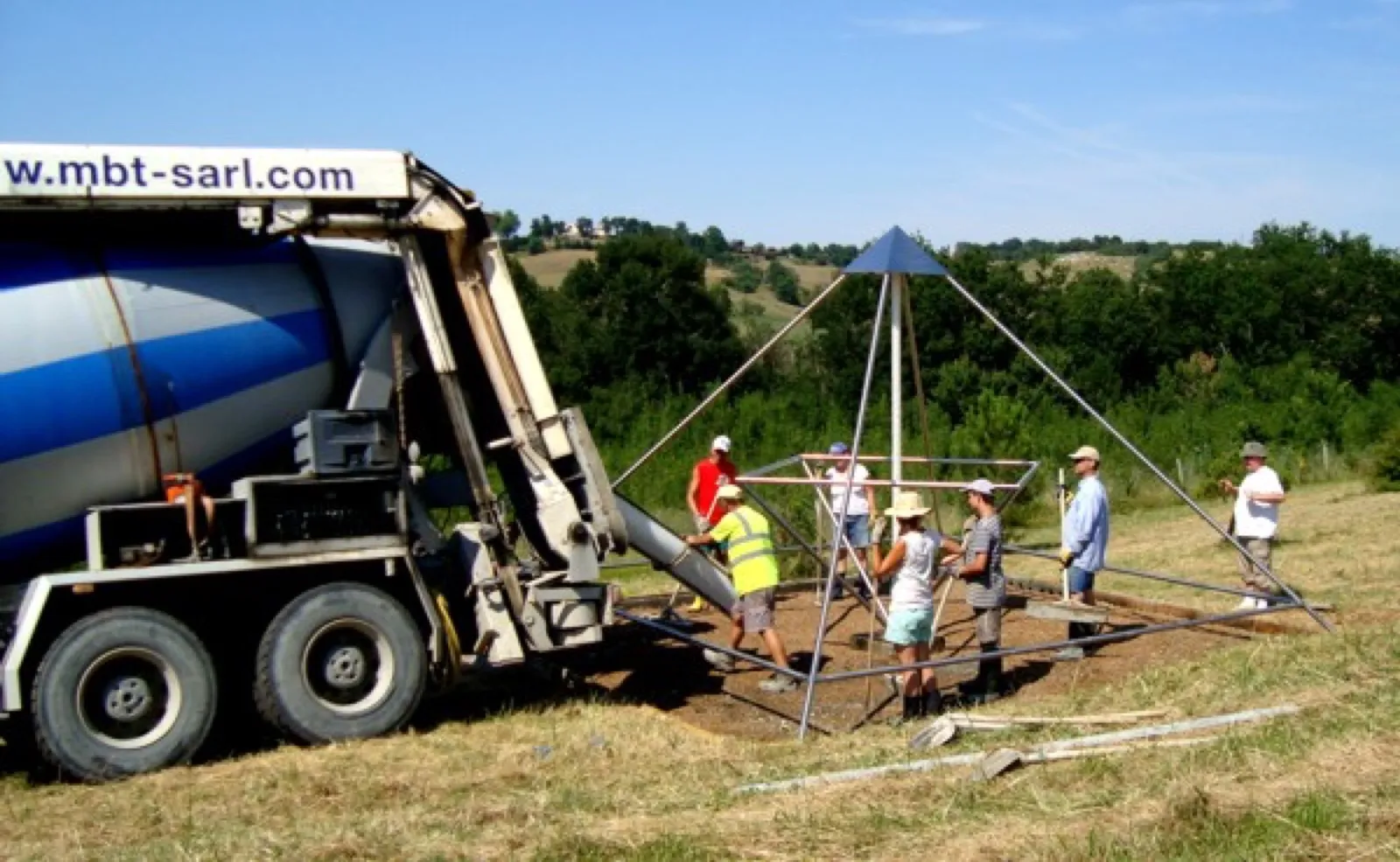 Baustelle auf einer sommerlichen Wiese: Ein Betonmischer-Lkw füllt über eine Rinne das Fundament unter einem Pyramidengestell mit blauer Spitze, mehrere Helfer mit Hüten, einer in gelber Warnweste, stehen daneben.
