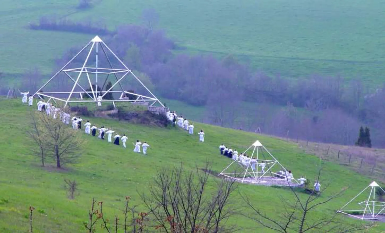 Eine lange Reihe weiß gekleideter Menschen zieht über einen grünen Hang an drei großen offenen Pyramidengestellen vorbei; weitere Gruppen stehen in und neben den Pyramiden. Im Hintergrund Wiesen, kahle Bäume und blühende Sträucher in dunstigem Licht.