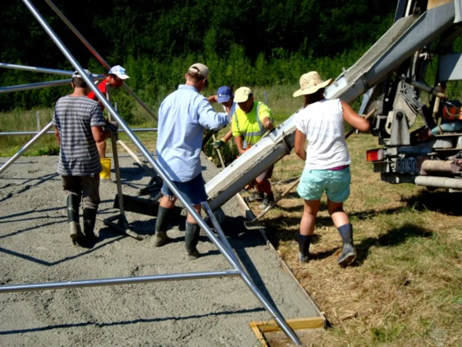 Mehrere Helfer in Gummistiefeln verteilen Beton, der über die Rutsche eines Betonmischfahrzeugs zwischen die Metallrohre eines Pyramidengestells fließt; im Hintergrund Wiese und dichter Wald.