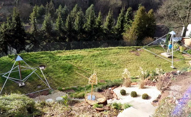 Blick von einer Steintreppe hinab in den Hanggarten: oben eine offene Energiepyramide mit innenliegendem Oktaeder im Rasen, davor die vergoldete Skulptur Vidaya auf einem Baumstumpf; rechts ein Kiesbeet mit drei kugelig geschnittenen Buchsbäumen.