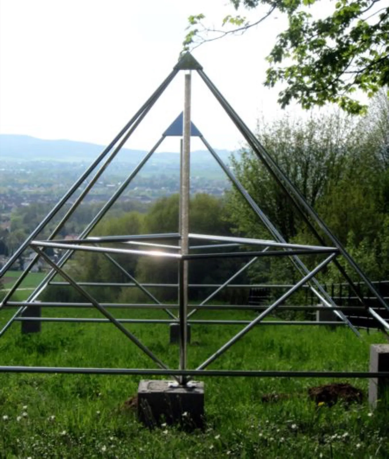 Zwei ineinander stehende offene Pyramiden aus Edelstahlrohren mit mittiger Achse auf Betonsockeln in einer Wiese; dahinter Bäume und ein weiter Blick über eine Stadt im Tal bis zu Hügeln.