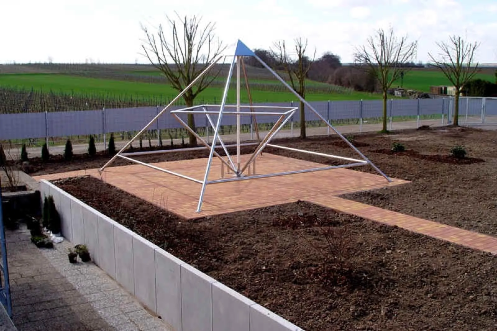 Energiepyramide aus Edelstahl mit blauer Spitze auf einem terrakottafarbenen Pflasterplatz inmitten frisch angelegter Erdbeete, gesäumt von einer Mauer aus Winkelsteinen; dahinter kahle Bäume, ein Zaun sowie Weinberge und Felder.
