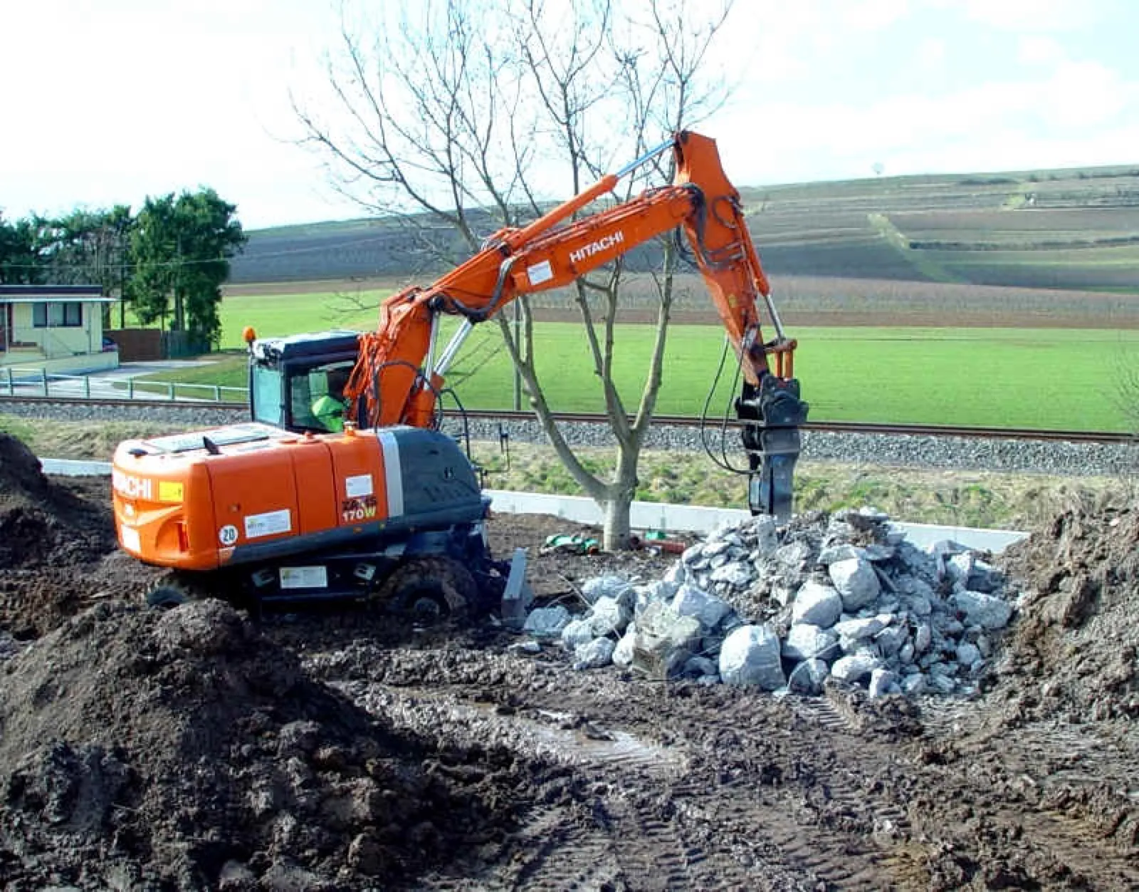 Ein orangefarbener Hitachi-Mobilbagger mit Hydraulikhammer bearbeitet einen Haufen Bruchsteine auf einer aufgewühlten Erdfläche; dahinter ein kahler Baum, Bahngleise, grüne Felder und Weinberge.