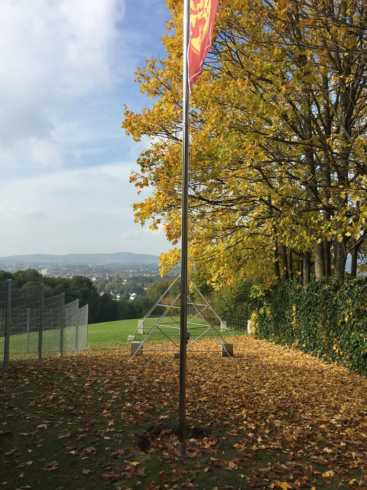 Hoher Metallmast mit rot-gelber Fahne auf laubbedecktem Rasen unter gelb gefärbten Herbstbäumen; dahinter eine offene Metallpyramide auf Betonsockeln, links ein Gitterzaun und ein weiter Blick über eine Stadt ins Tal.