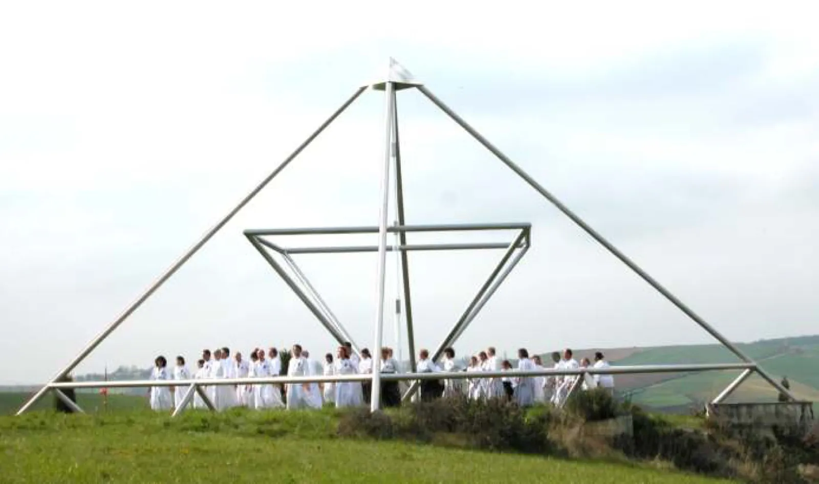 Große offene Pyramide aus Metallrohren mit innen verdreht eingesetztem Rahmen auf einer Wiesenkuppe; darin steht eine Gruppe von rund dreißig Menschen in weißer Kleidung, dahinter Felder unter bedecktem Himmel.