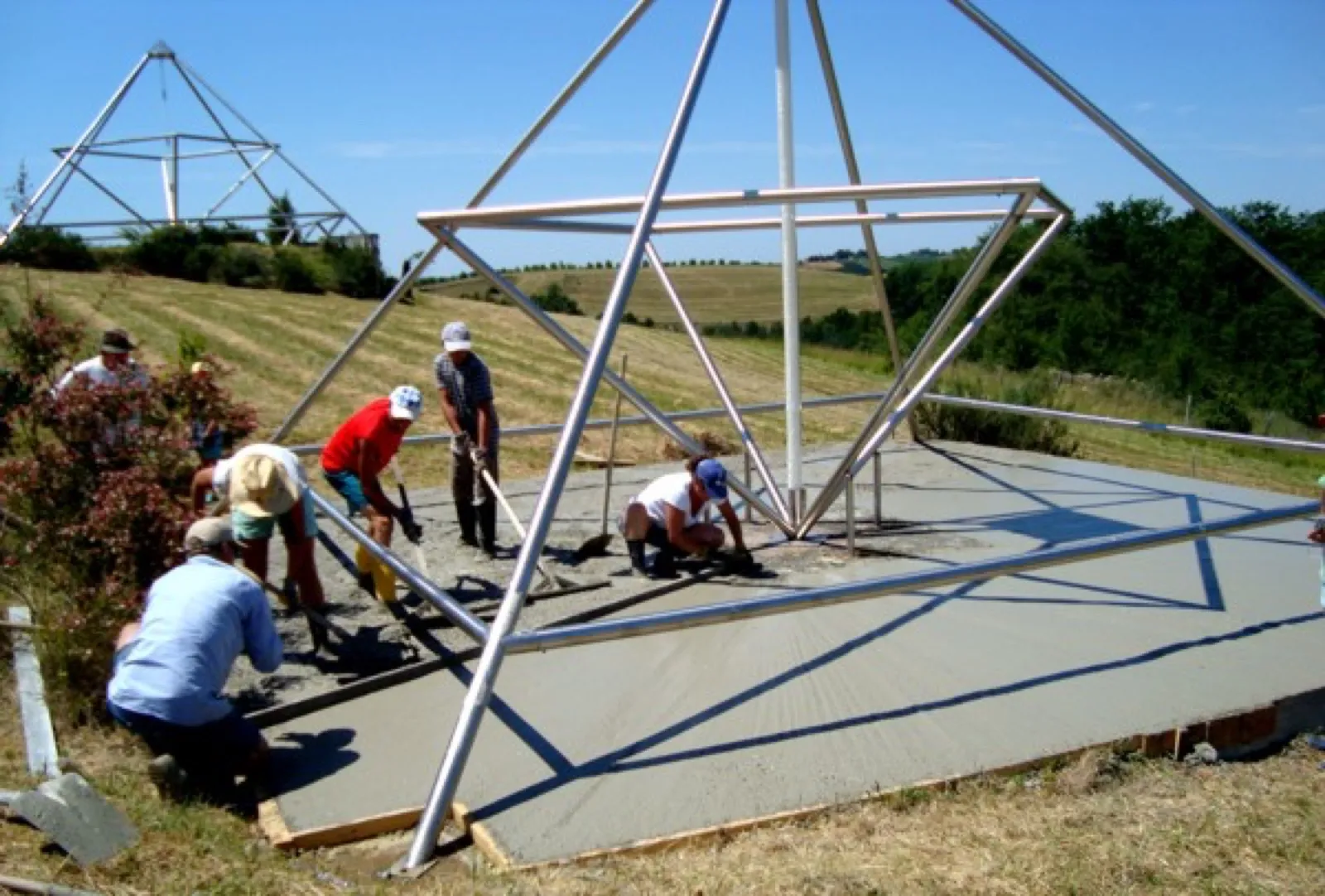 Mehrere Helfer mit Hüten und Kappen glätten mit Schaufeln und Rechen eine frisch gegossene Betonfläche unter einem großen offenen Pyramidengestell. Im Hintergrund eine zweite Pyramide auf einer Anhöhe, Getreidefelder und Bäume.