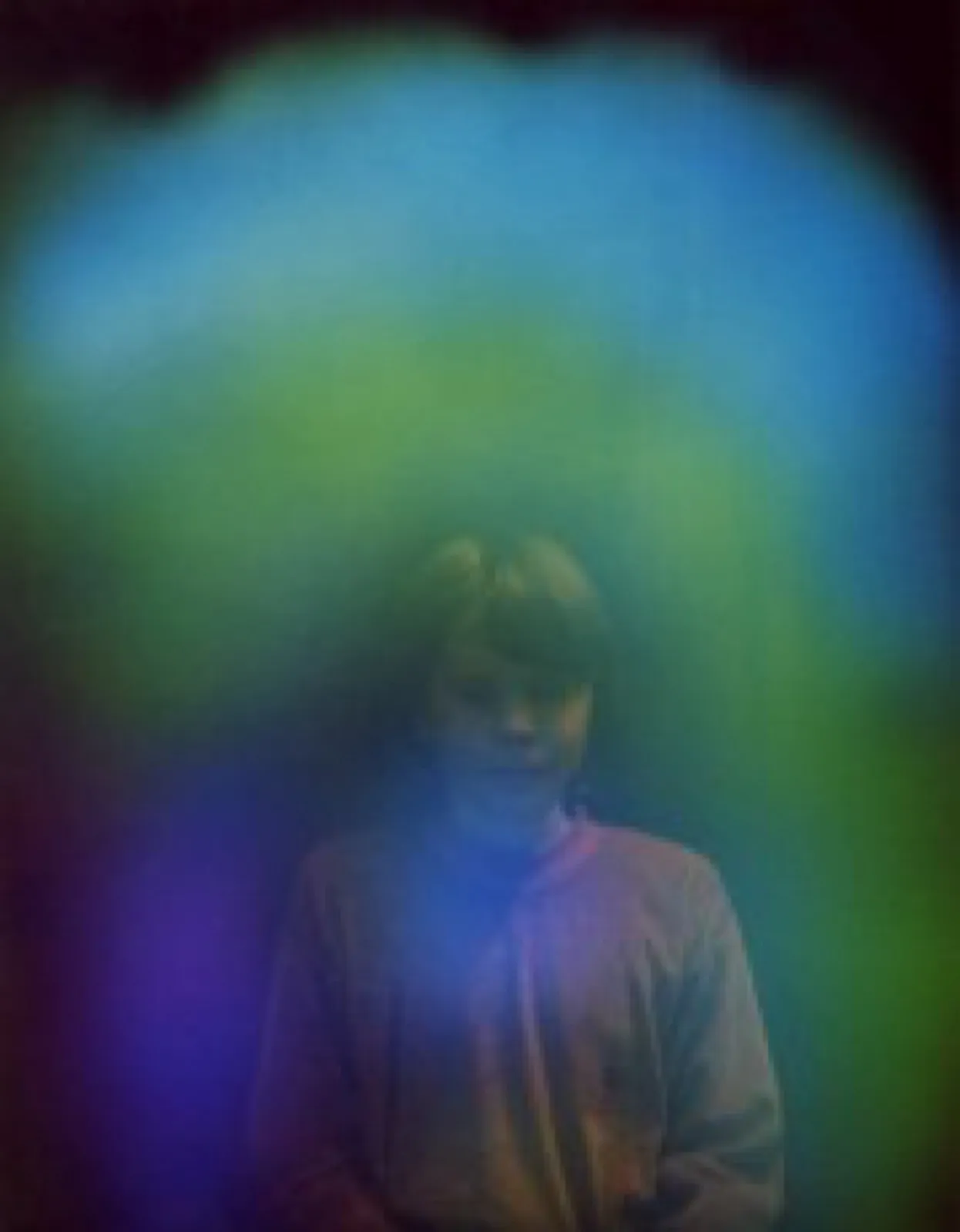 Aurafoto einer jungen Person mit halblangem Haar in hellem Oberteil vor dunklem Hintergrund. Ein blau-grüner Farbschleier liegt über Kopf und Schultern, unten geht er in Violett über.