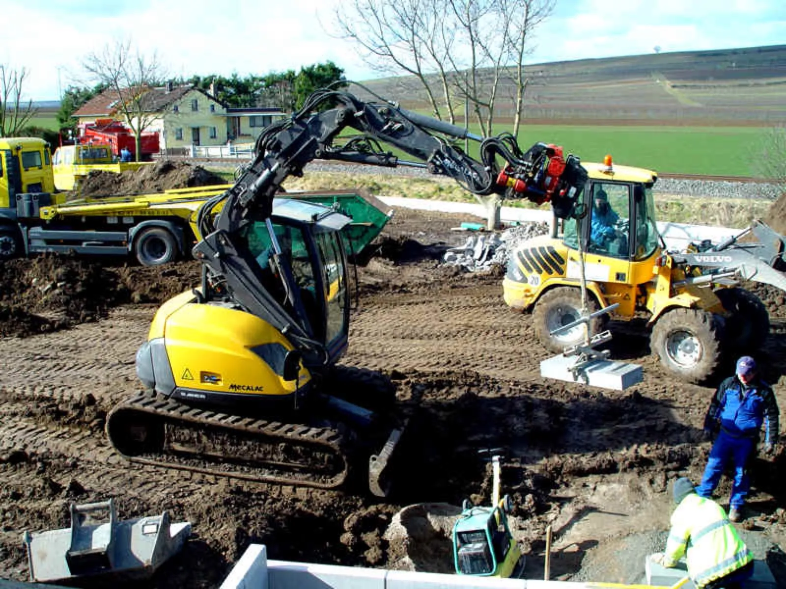 Baustelle mit einem gelben Kettenbagger, einem gelben Radlader, der eine Betonplatte am Haken hält, und einem Lkw mit Container; Arbeiter in Arbeitskleidung und Warnjacke stehen daneben. Im Hintergrund ein Wohnhaus, Bäume und Weinberge.