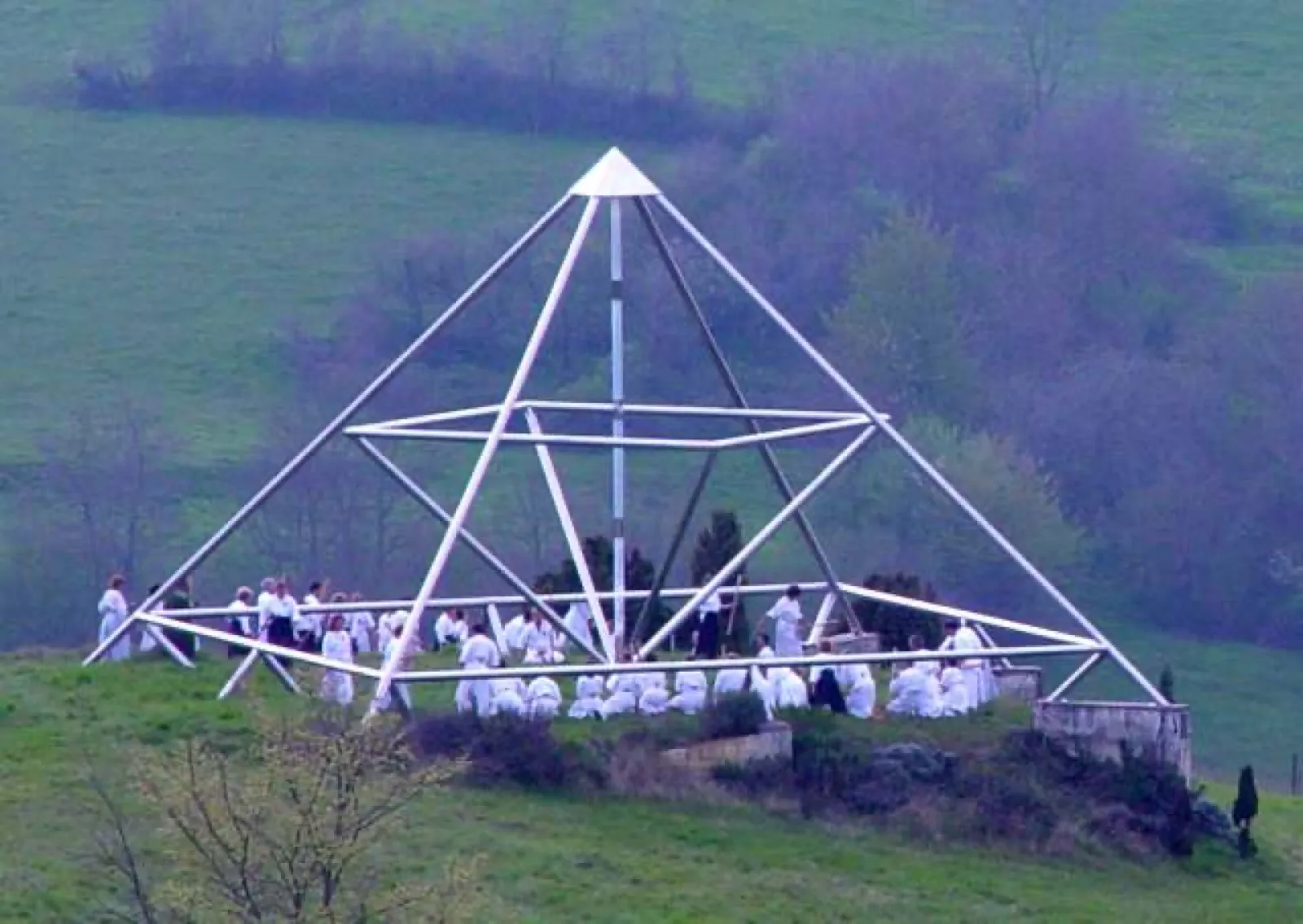 Große offene Energiepyramide aus Edelstahlrohren mit Mittelachse und innerem Rahmen auf einer Wiese; darin sitzen und stehen zahlreiche weiß gekleidete Menschen, rechts ein niedriger Mauerrest, dahinter bewaldete Hügel.
