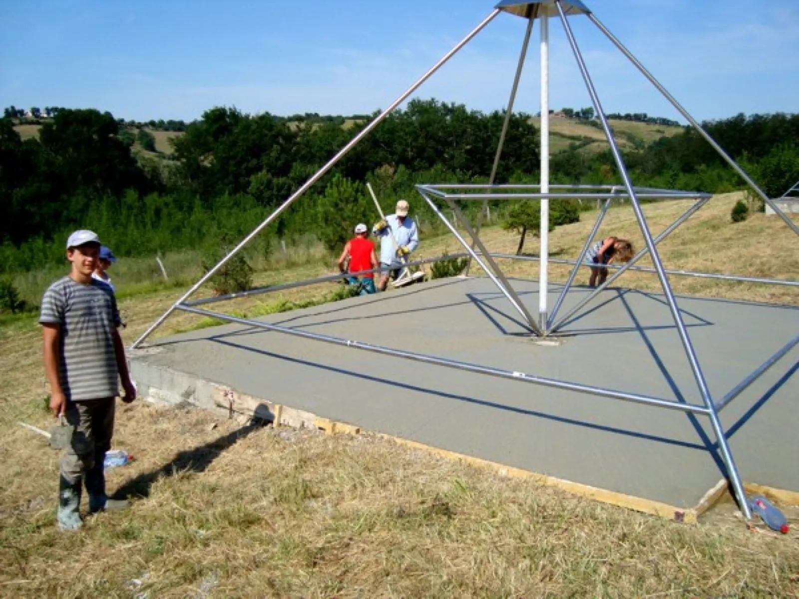 Frisch gegossene Betonplatte unter einer großen offenen Pyramide aus Metallrohren mit mittiger Achse; mehrere Helfer arbeiten am Rand, vorn links steht ein Junge in gestreiftem T-Shirt und Gummistiefeln, dahinter bewaldete Hügel.