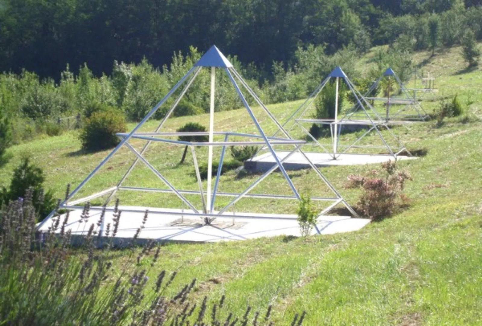 Drei offene Pyramiden aus Edelstahlrohren mit blauen Spitzen stehen gestaffelt auf hellen Betonplatten an einem grünen Hang, im Vordergrund blühender Lavendel, dahinter Waldrand.