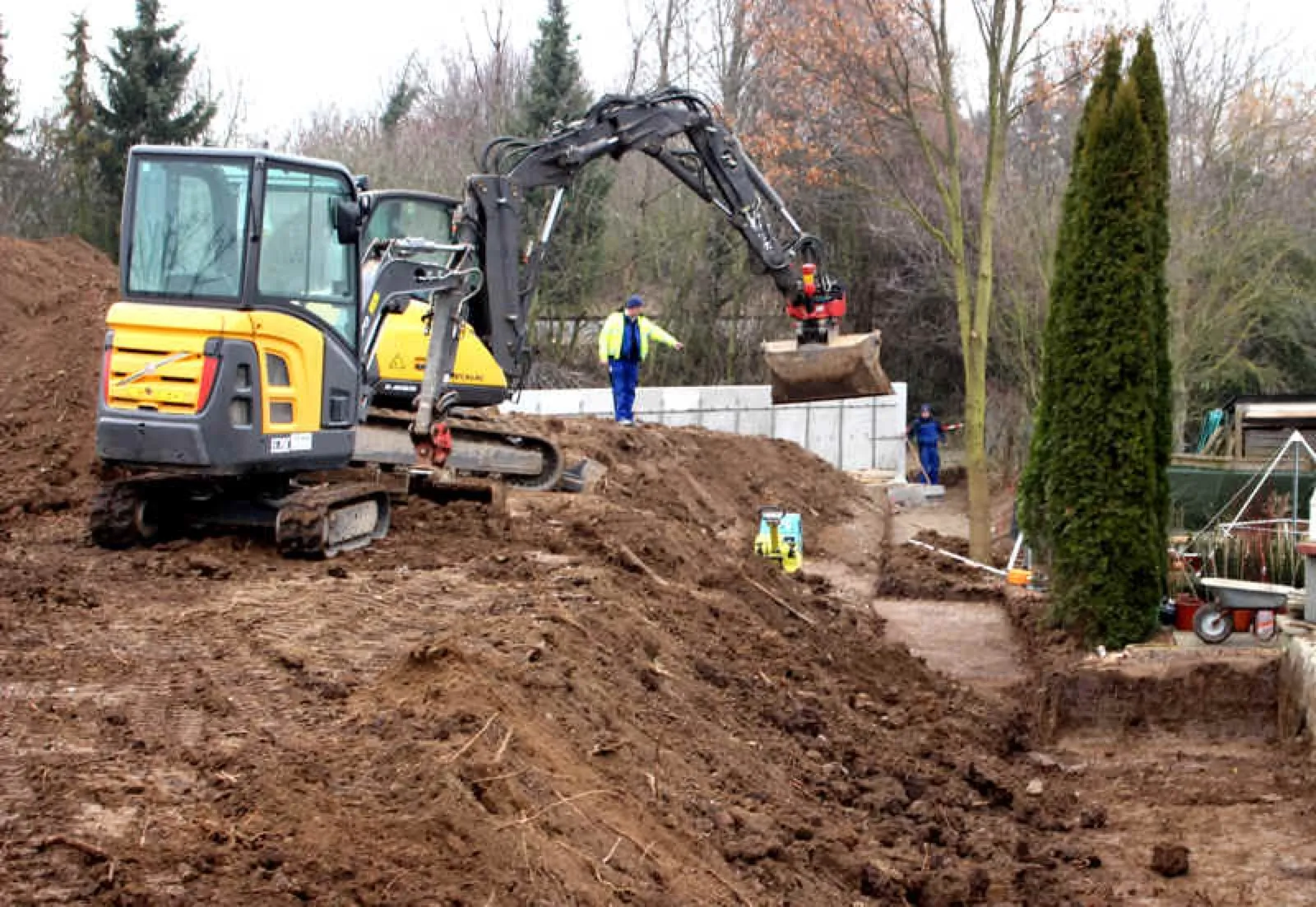 Bauphase im Garten des Kyborg Instituts: Ein gelber Minibagger versetzt Erde, zwei Arbeiter in Blau mit Warnweste stehen an einer Reihe Betonwinkelsteine; rechts eine Zypresse und am Bildrand ein Teil eines Pyramidengestells.