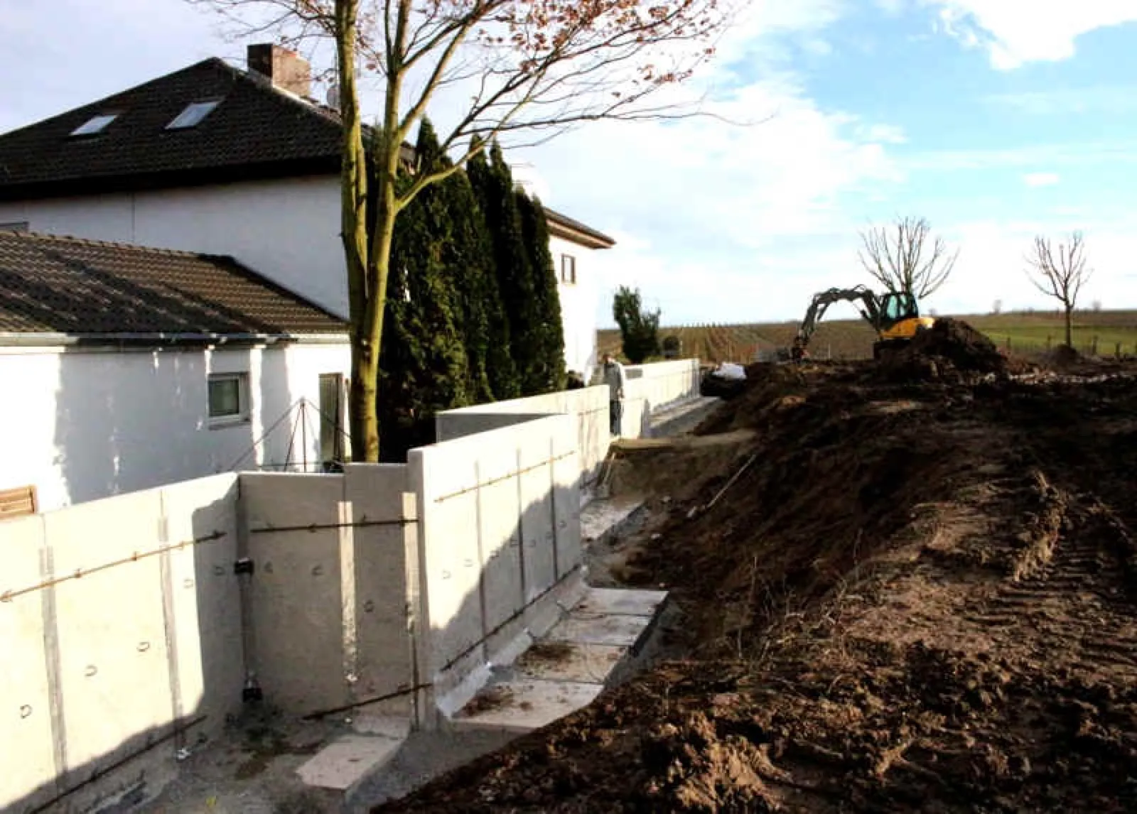 Neu gesetzte Betonstützmauern entlang eines Wohnhauses mit Dachfenstern, daneben ein Graben und aufgeschüttete Erde; im Hintergrund ein kleiner Bagger, kahle Bäume und Weinberge.