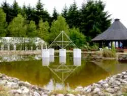 Energiepyramide Modell C mit weißen Platten an einem Teich, im Wasser gespiegelt, mit Pavillon im Hintergrund (Golfplatz im Taunus)