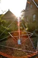 Energiepyramide Modell C in einem Garten vor einem Backsteinhaus, mit Sonnenstern im Gegenlicht