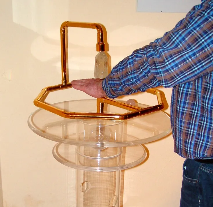 Eine Person hält die Hände flach über den Brunnen von Atlantis; der achteckige Kupferring ruht auf Acrylplatten auf einer Holzsäule