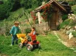 Zwei Kinder spielen mit Tret-Fahrzeugen vor einer rustikalen Berghütte; im Hintergrund eine Horus-Energiepyramide Modell C