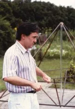 Ein Mann mit Ruten/Drähten im Garten, dahinter ein frühes Modell C
