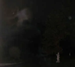 Dunkle Nachtaufnahme eines Parks mit Baum und kleiner Statue; links oben eine nebelhafte helle Erscheinung