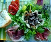 Ein frischer grüner Salat mit Geflügelmägen (Salade de gésiers), Rohschinken, Tomaten und Weißbrot auf einem Glasteller.