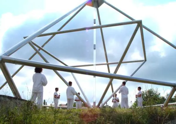 Die große 9-Meter-Energiepyramide aus Metallstreben im Freien unter wolkigem Himmel; eine Gruppe weiß gekleideter Menschen meditiert rund um die senkrechte Mittelachse.