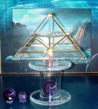 Energiepyramide Modell A – eine kleine goldene Doppelpyramide – auf einer durchsichtigen Acrylsäule, umgeben von violetten Amethyst-Kristallkugeln, vor einem Landschaftsbild.