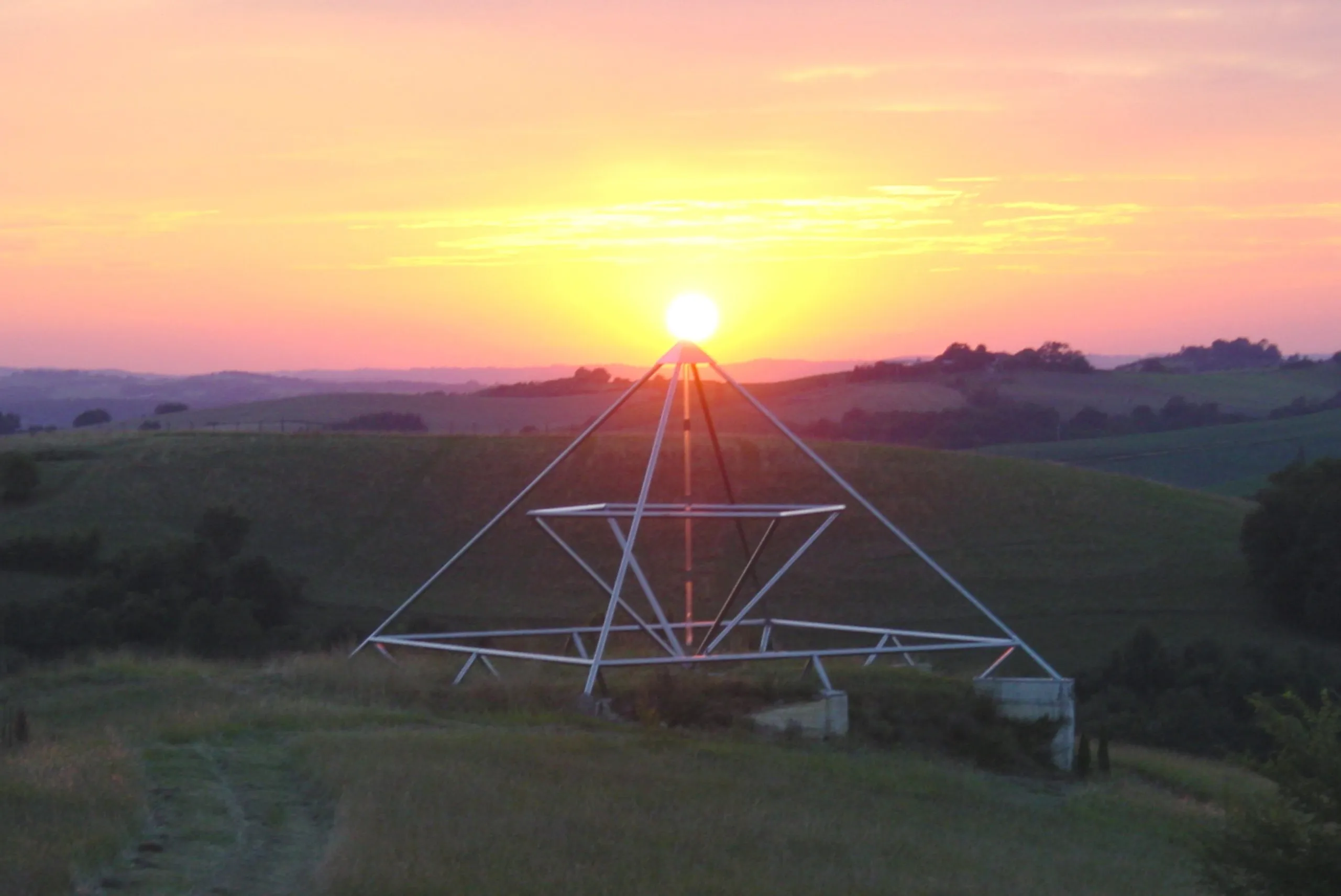 Die große Energiepyramide Modell G als Metallrahmen-Doppelpyramide im Gegenlicht eines Sonnenuntergangs über sanften Hügeln; die Sonne steht genau an der Spitze.