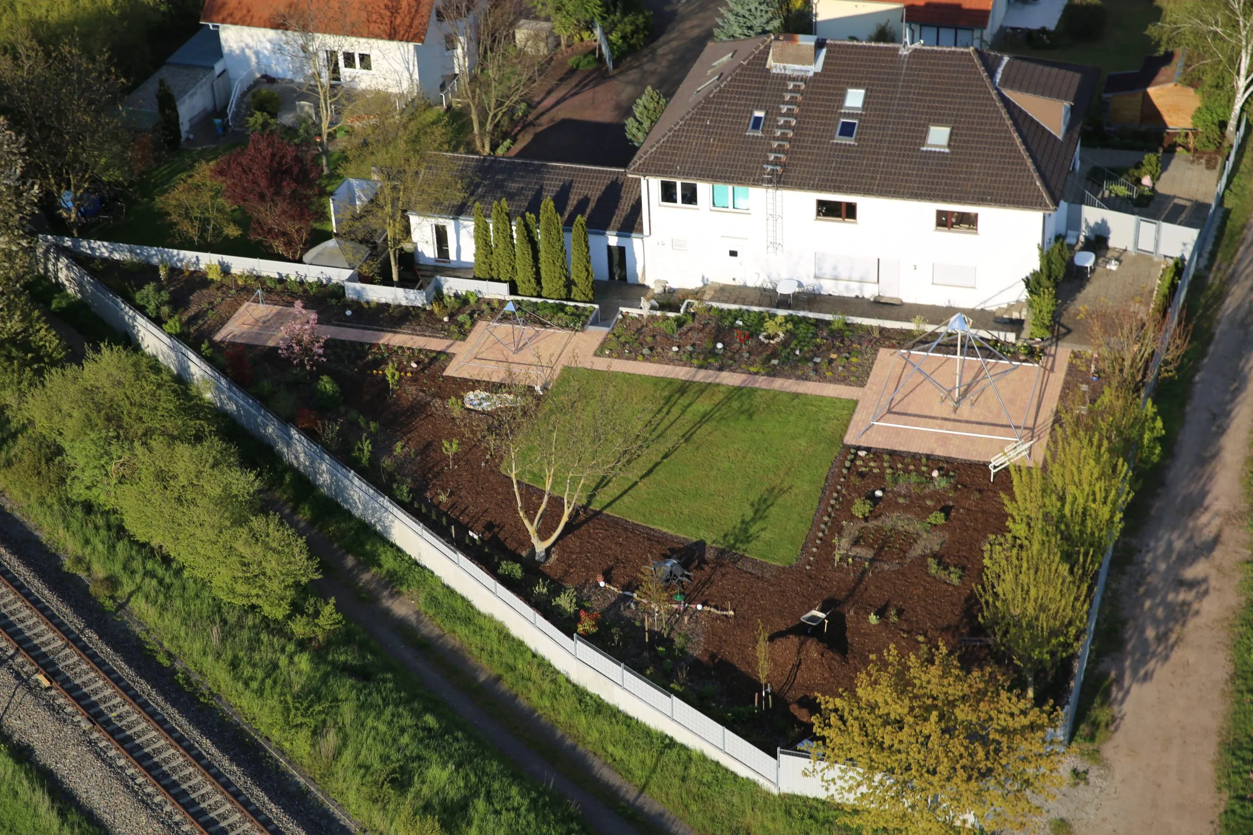 Luftbild des Kyborg Instituts in Flörsheim-Dalsheim: weißes Wohnhaus mit dunklem Dach und ummauertem Garten mit Wegen, Beeten und einer Rasenfläche, auf der das Gerüst einer Horus Energiepyramide steht; links unten verläuft eine Bahnlinie