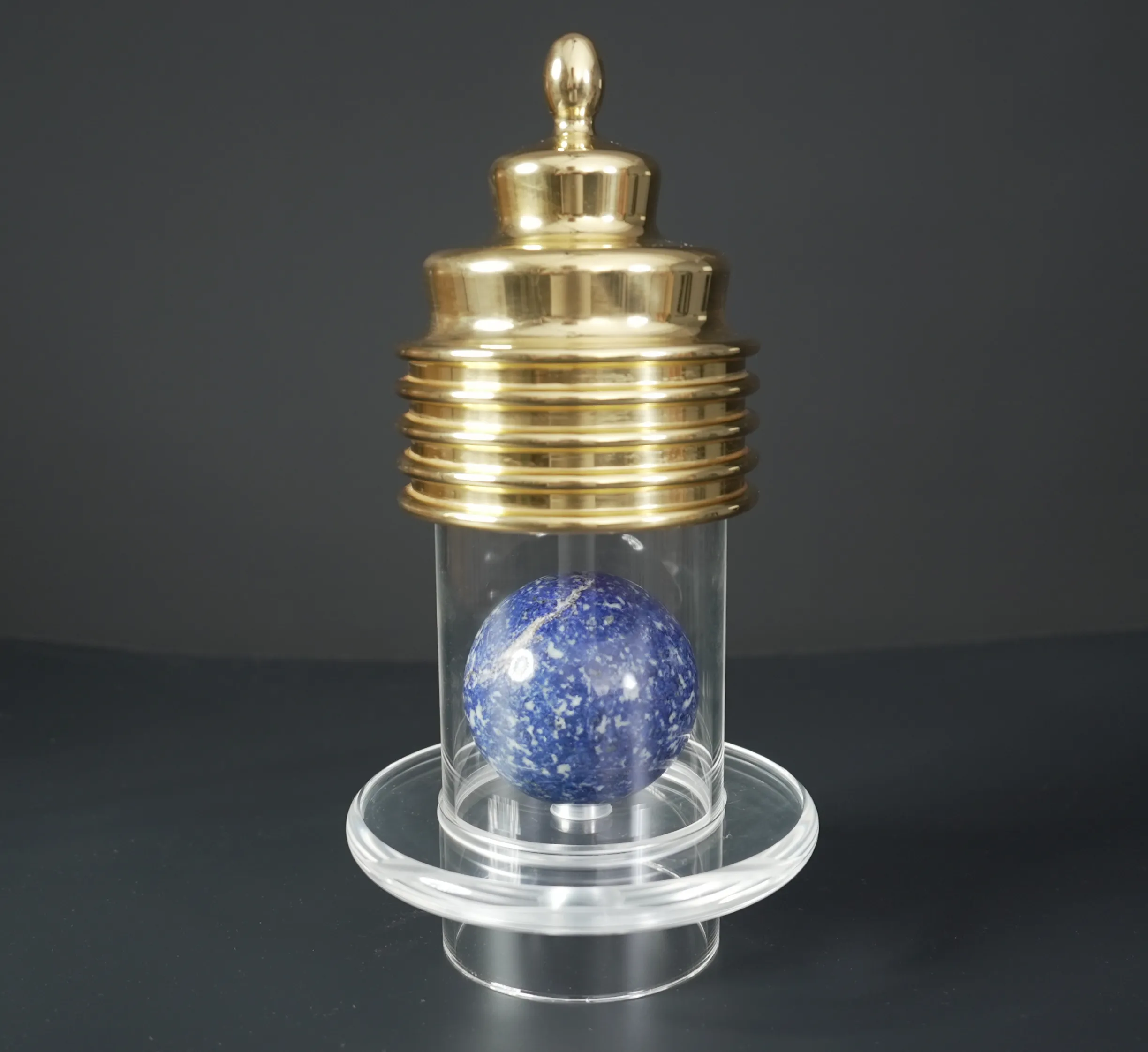 Die goldene Mega Power Cap auf einer Acrylsäule mit blauer Lapislazuli-Kugel vor schwarzem Hintergrund