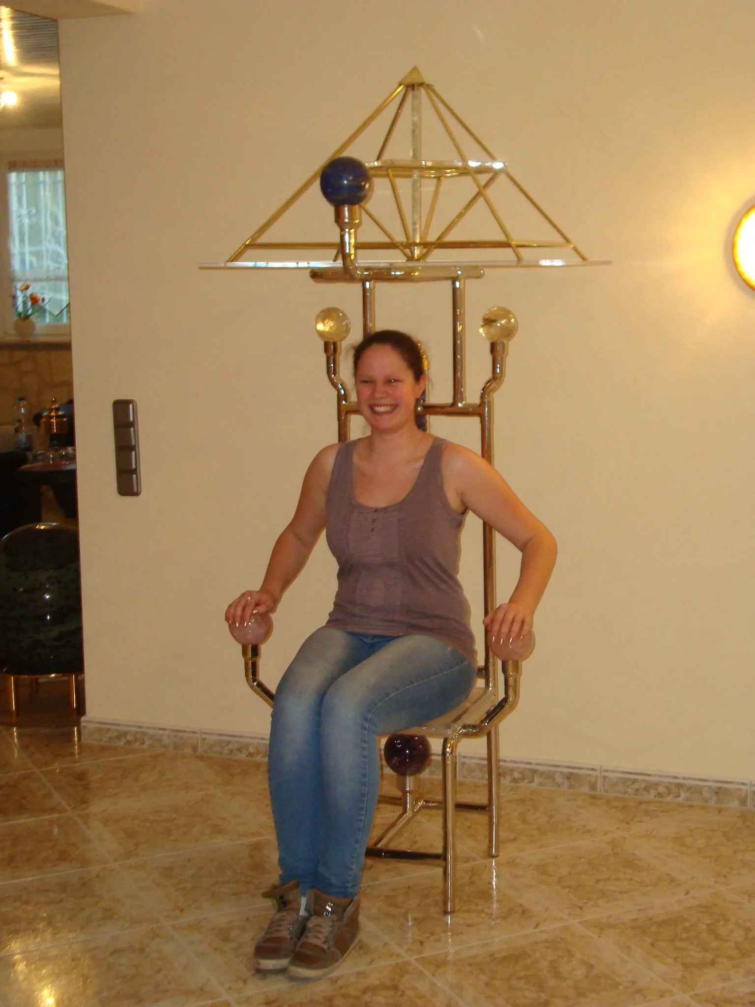 Eine junge Frau sitzt auf dem Horus-Pharaonenstuhl mit großer goldener Energiepyramide Modell B und farbigen Kugeln an den Armlehnen