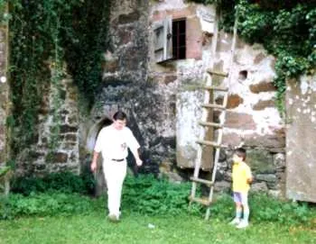 D. Harald Alke, weiß gekleidet, geht Hand in Hand mit einem kleinen Kind an einer alten, verwitterten Bruchsteinmauer entlang; rechts lehnt eine Holzleiter.