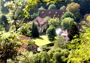 Blick aus dem Wald auf eine Klosteranlage mit Fachwerkbauten, eingebettet in ein grünes Tal – das Schloss/Kloster Schönbrunn aus der Ferne.