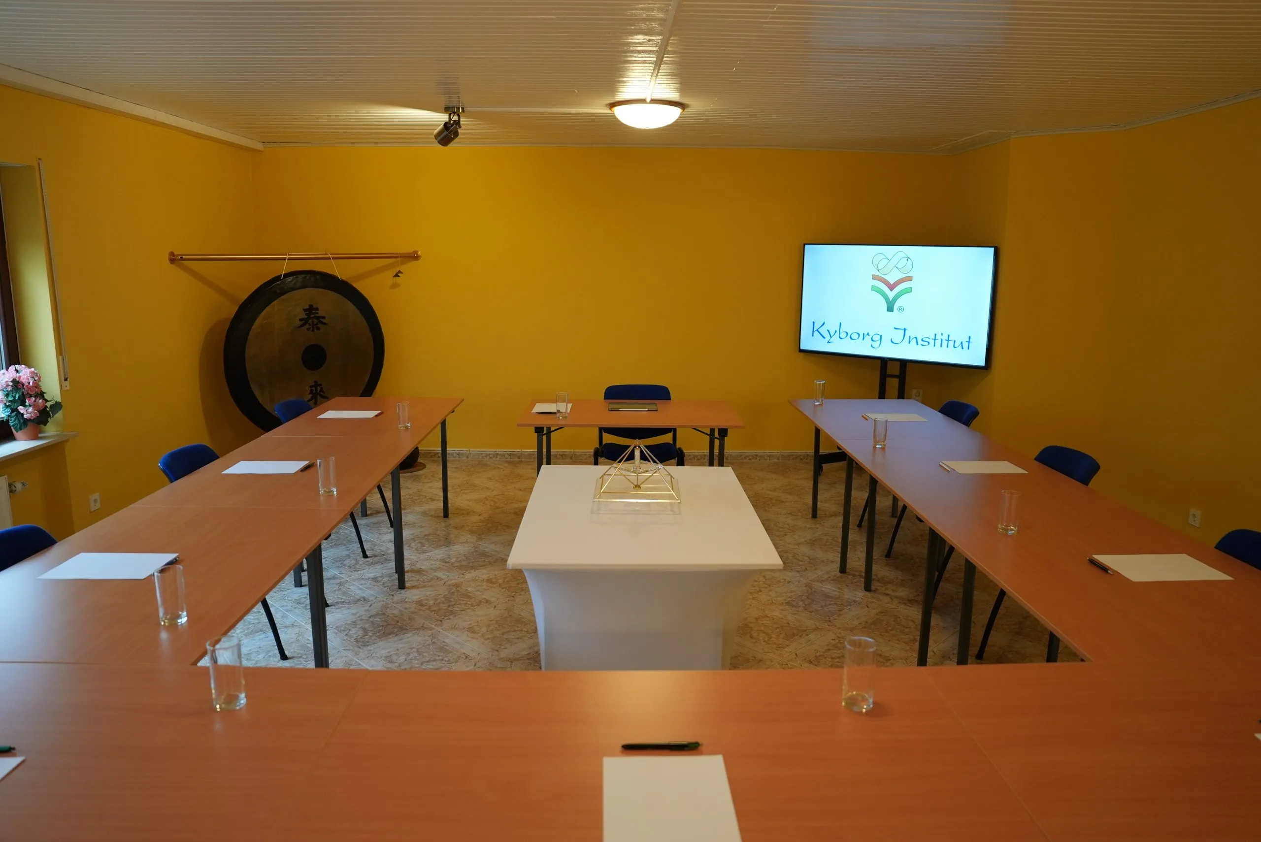 Seminarraum des Kyborg Instituts mit U-förmig gestellten Holztischen und blauen Stühlen, einem großen Gong an der Wand, einem Bildschirm mit dem Kyborg-Institut-Logo und einem kleinen goldenen Pyramidenmodell auf dem Mitteltisch