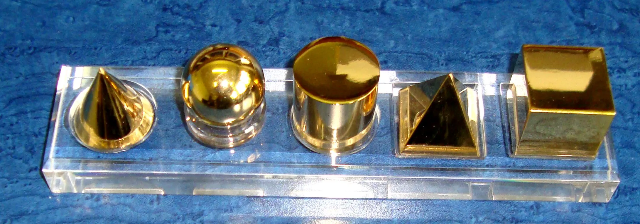 Der Satz des Archimedes: fünf vergoldete Körper (Kegel, Halbkugel, Zylinder, Pyramide, Würfel) auf einer Acrylleiste vor blauem Hintergrund